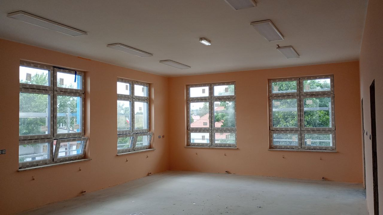 Świeżo pomalowane wnętrze biura z kilkoma oknami i zamontowanym oświetleniem sufitowym. Ściany w odcieniu brzoskwiniowym, surowa podłoga betonowa, okna częściowo otwarte.