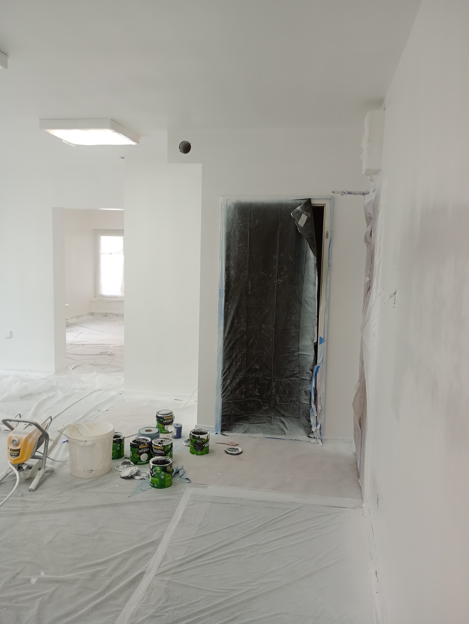 Przygotowania do malowania: pomieszczenie zabezpieczone folią malarską, puszki z farbą Bondex Ceramic i agregat malarski na podłodze, drzwi oklejone folią ochronną, świeżo pomalowane ściany na biało.