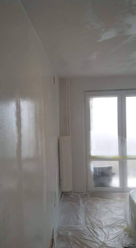 Świeżo pomalowana ściana w pokoju, z widoczną mokrą fakturą farby, okno zabezpieczone folią malarską, podłoga również przykryta folią ochronną.