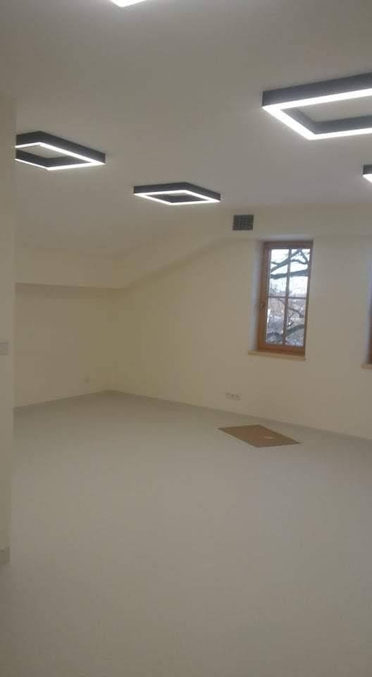 Pomalowane na jasny odcień wnętrze poddasza z oknem w drewnianej ramie i nowoczesnym oświetleniem sufitowym w geometryczne kształty.