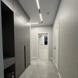 Nowoczesny korytarz z szarą zabudową szaf, podświetleniem LED w suficie i płytkami podłogowymi w odcieniach szarości, widok na otwarte drzwi do kolejnych pomieszczeń.