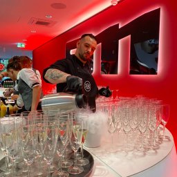 Jemy Event Space MILENA BACHOWSKA - Barman w czarnym stroju, z tatuażami, nalewa dymiący napój do kieliszków na tacy. Czerwone oświetlenie, nowoczesny bar.