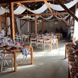 Jemy Event Space MILENA BACHOWSKA - Rustykalna sala weselna w stodole z drewnianymi stołami, białymi krzesłami, kwiatami i bufetem. Dekoracje z białego materiału pod sufitem.