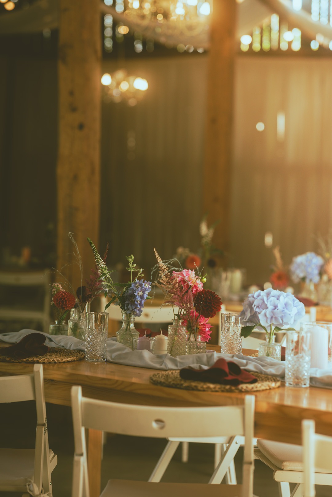 Dekoracja stołu z kwiatami w szklanych wazonach, świecami i serwetkami na drewnianym stole z białymi krzesłami w tle, oświetlona ciepłym światłem.