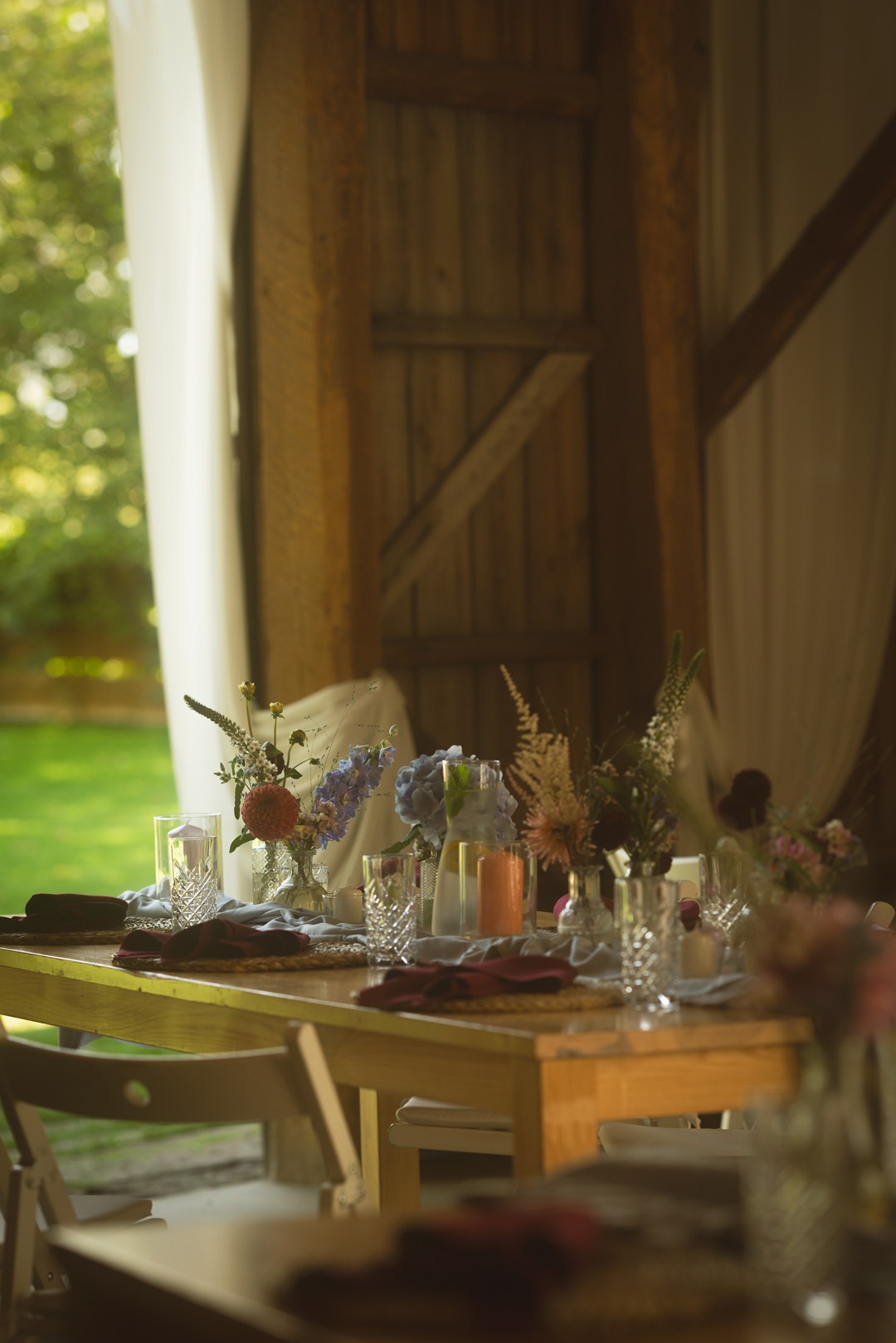 Rustykalny stół z dekoracjami kwiatowymi i szklanymi naczyniami, ustawiony w drewnianej stodole z widokiem na zieleń, tworzący elegancką aranżację.