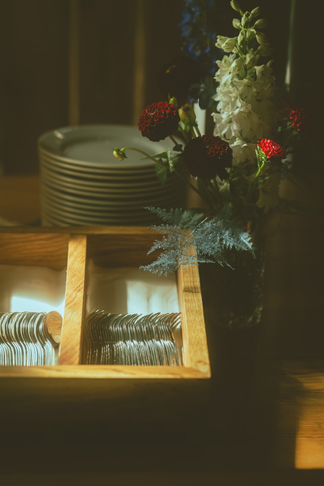 Stół z drewnianą skrzynką pełną sztućców, stosem talerzy i wazonem z kwiatami w odcieniach czerwieni, bieli i niebieskiego, oświetlony ciepłym światłem.