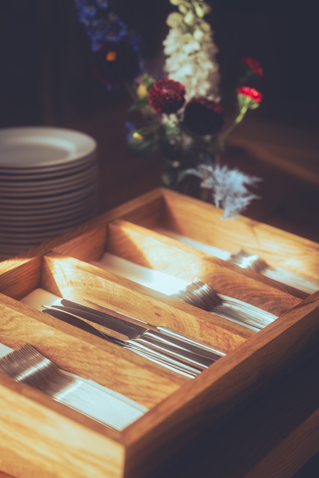 Drewniany organizer na sztućce z nożami, widelcami i łyżkami, w tle stos talerzy i bukiet kwiatów. Przygotowanie do eleganckiego przyjęcia.