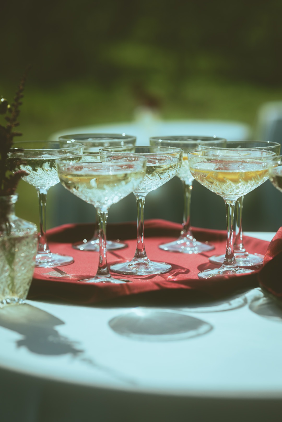 Kryształowe kieliszki do szampana wypełnione napojem, ustawione na czerwonej tacy, z dekoracyjnym wazonem i serwetką, w plenerze.