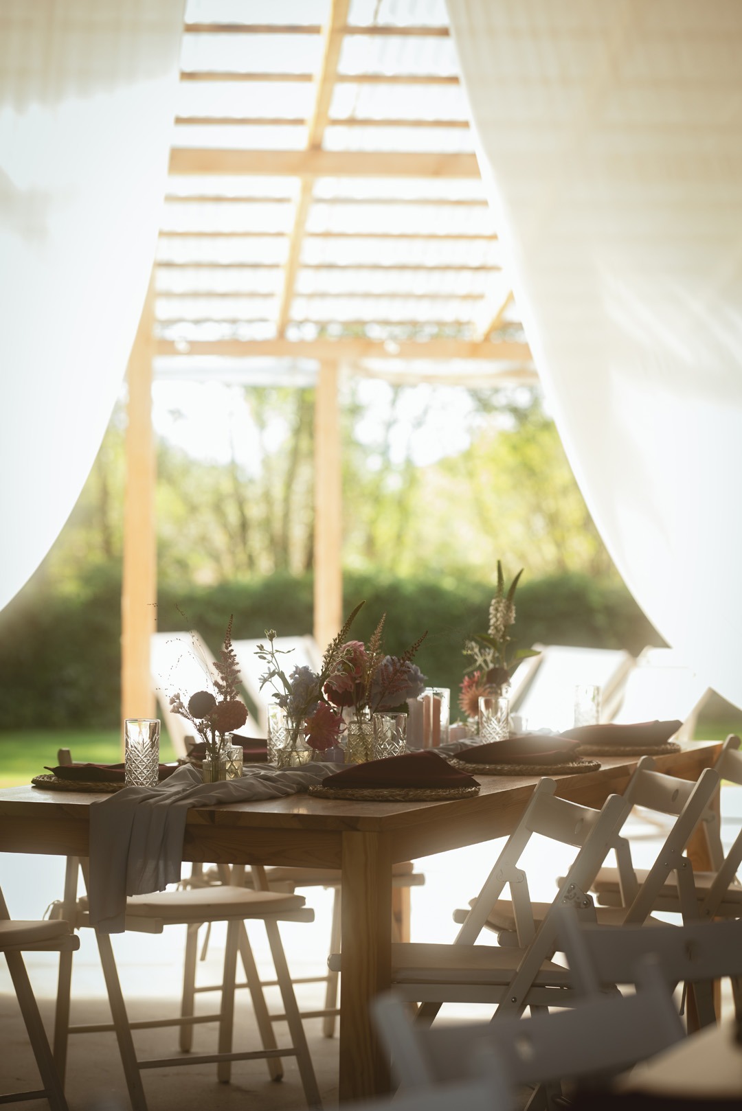 Elegancko nakryty stół z kwiatami w szklanych wazonach, brązowe podkładki i szara serweta, ustawiony na zewnątrz pod drewnianą konstrukcją z białymi zasłonami.