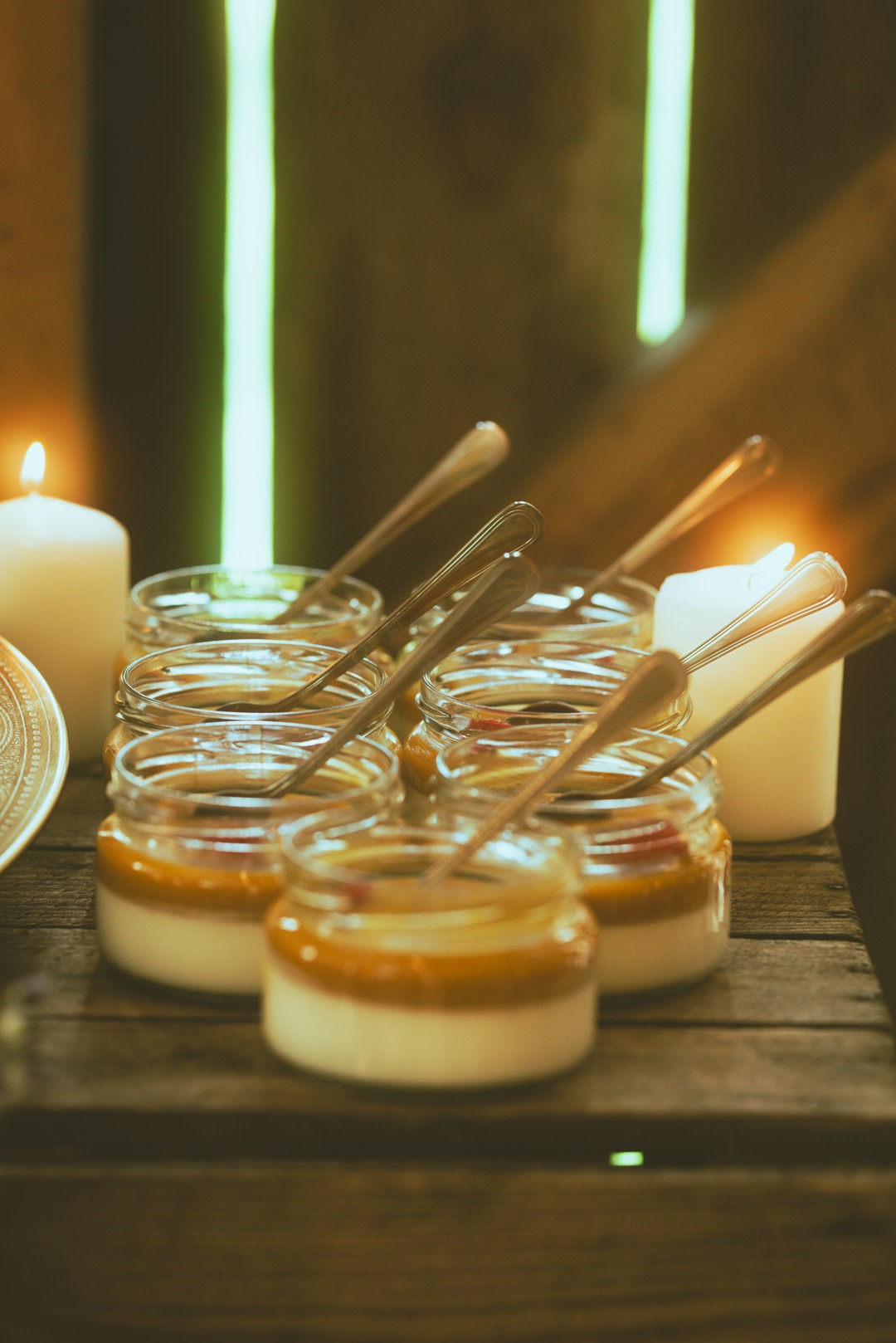Deser w słoiczkach z łyżeczkami, otoczony świecami, na drewnianym stole. Ciepłe światło tworzy przytulną atmosferę.