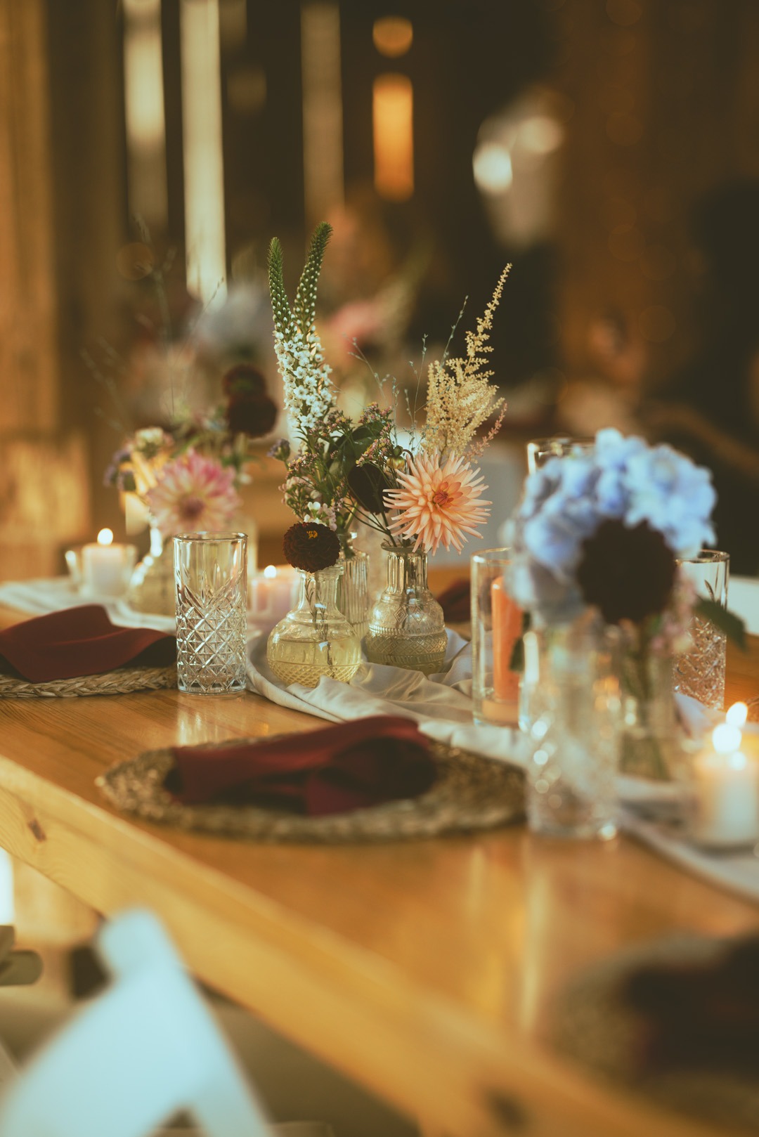 Dekoracja stołu z kwiatami w szklanych wazonach i świecami, nakrycie z bordowymi serwetkami na drewnianym stole, ciepłe oświetlenie, elegancki wystrój.