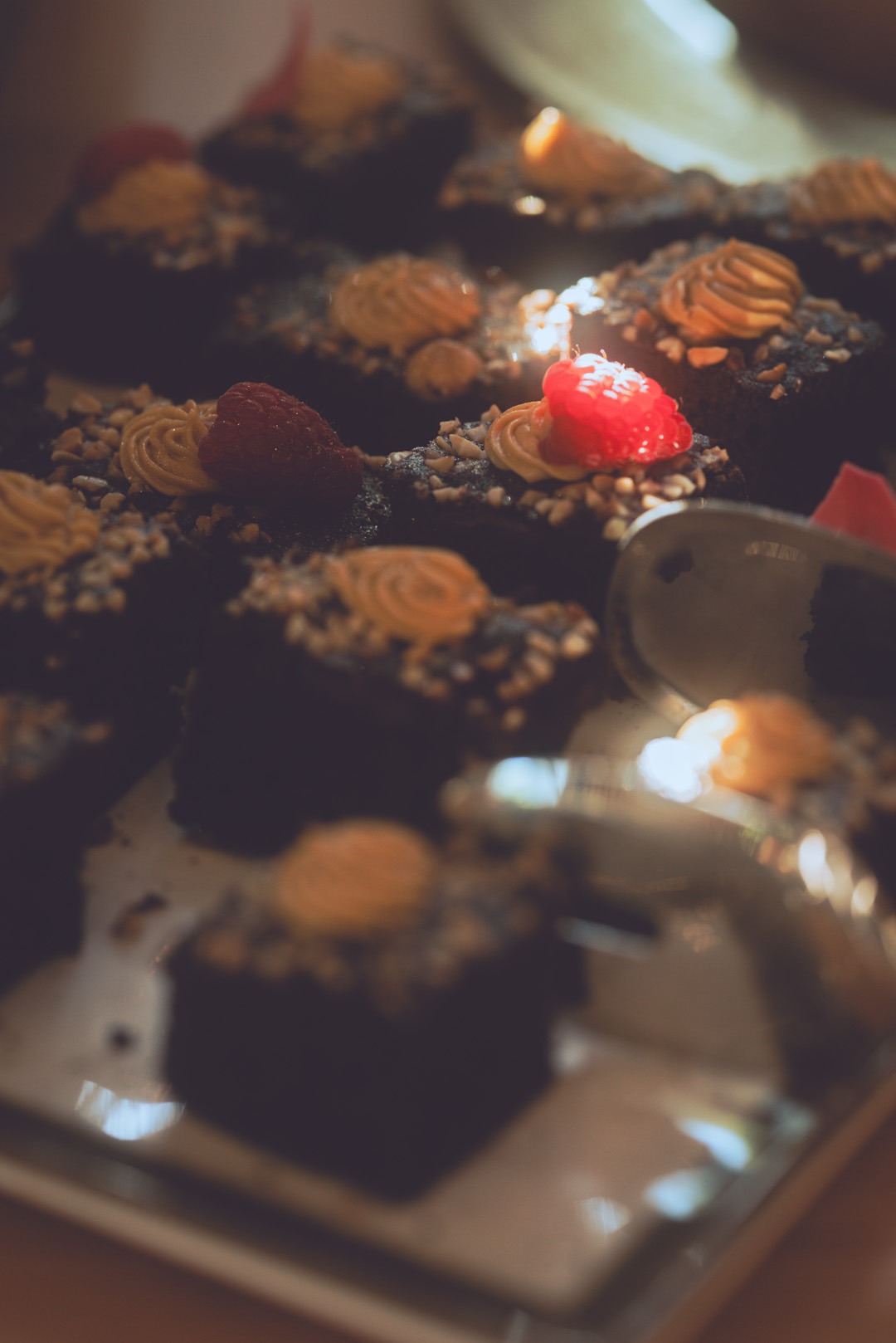 Ciemne babeczki z kremem, posypką orzechową i malinami na srebrnej tacy, oświetlone ciepłym światłem. Dekoracja z owocami i kremem.