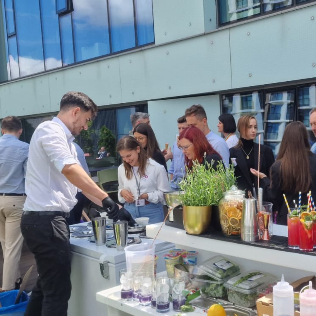 Barman w akcji, przygotowuje kolorowe drinki na świeżym powietrzu dla grupy osób podczas eventu firmowego. Widoczne składniki, naczynia barmańskie i dekoracje.