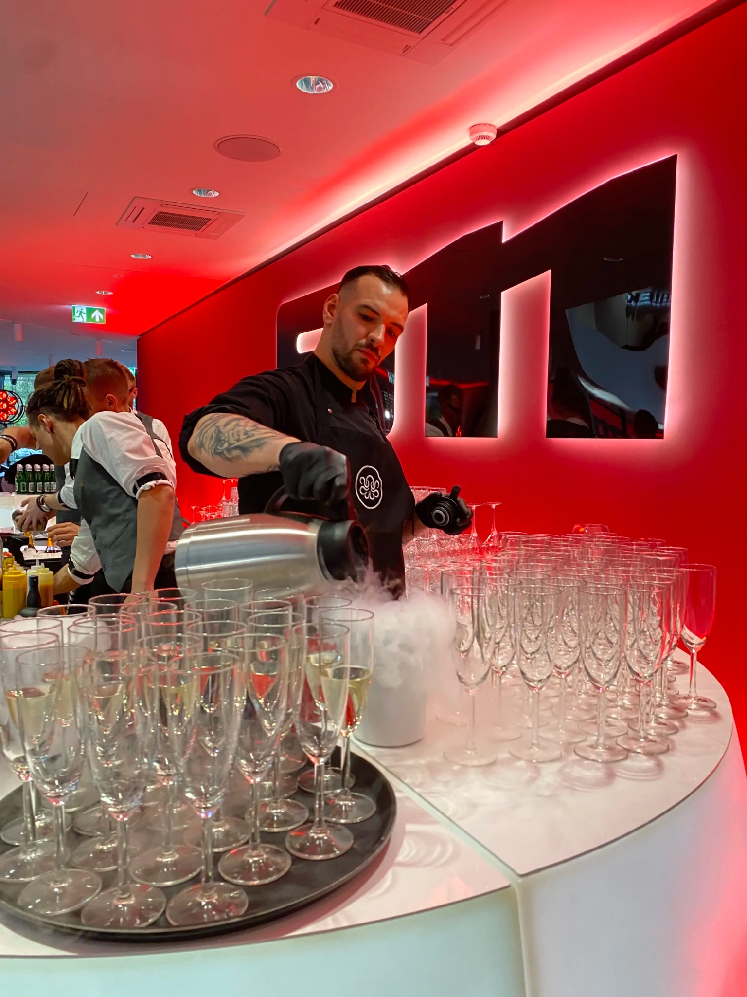 Barman w czarnym stroju, z tatuażami, nalewa dymiący napój do kieliszków na tacy. Czerwone oświetlenie, nowoczesny bar.