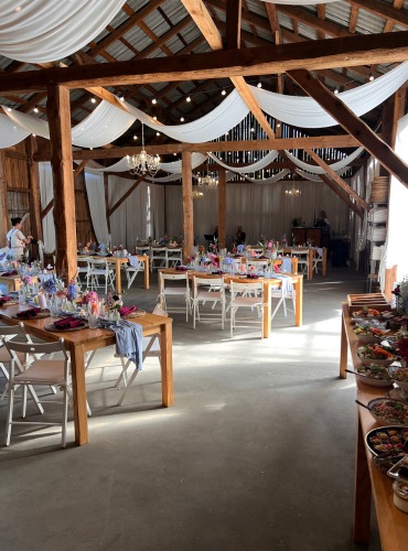 Rustykalna sala weselna w stodole z drewnianymi stołami, białymi krzesłami, kwiatami i bufetem. Dekoracje z białego materiału pod sufitem.