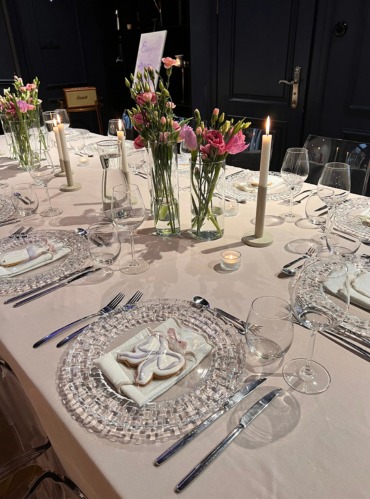 Elegancko nakryty stół z kwiatami, świecami i dekoracyjnymi ciasteczkami na przezroczystych talerzach. Subtelne oświetlenie tworzy intymną atmosferę.