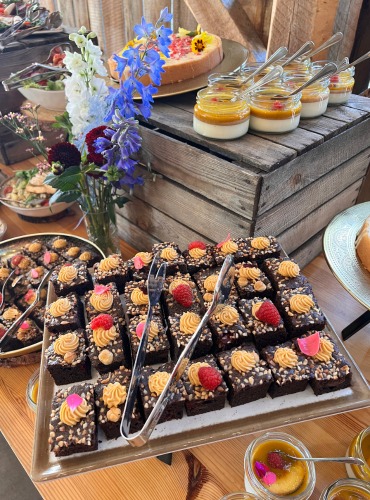 Bogato zastawiony stół z deserami: mini brownie z malinami, kremowe desery w słoiczkach, ciasto z kwiatami i sałatki. Dekoracja kwiatowa w wazonie.