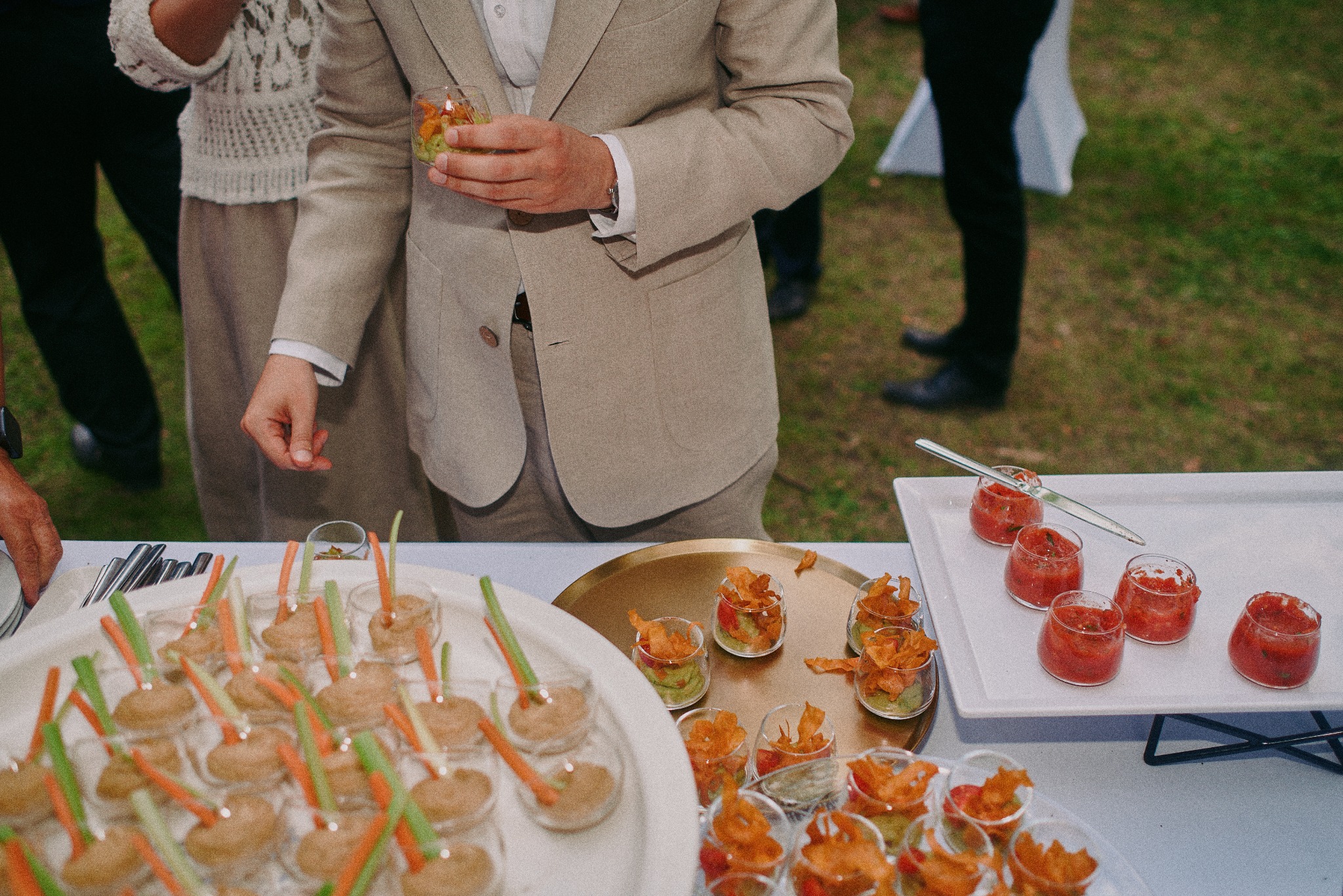 Elegancki catering: osoba w beżowym garniturze trzyma kieliszek z przekąską. Na stole tace z mini-przystawkami w szklanych naczyniach, widok z góry, przyjęcie w plenerze.
