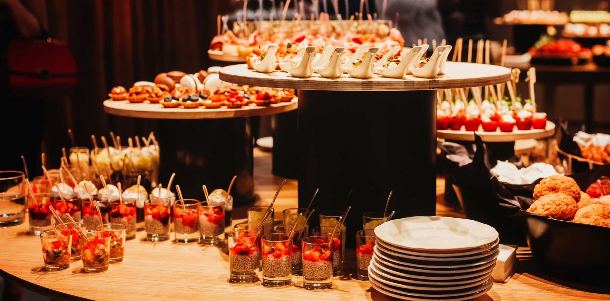 Bogato zastawiony stół z różnorodnymi przekąskami: mini desery w szklankach, tartaletki z owocami, kanapeczki i inne smakołyki. Idealne na catering imprezowy.