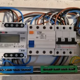 Rozdzielnica elektryczna z licznikiem energii, bezpiecznikami i przewodami w kolorach niebieskim, brązowym, czarnym, szarym i żółto-zielonym, zamontowana w białej obudowie na jasnej ścianie.