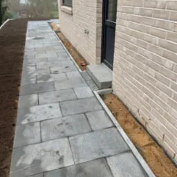 Świeżo ułożona, szara kostka brukowa prowadząca do wejścia domu z jasnej cegły, z widocznym obrzeżem i przygotowanym terenem pod trawnik.