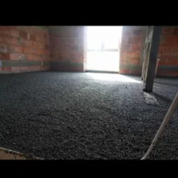 Świeżo wylana, ciemna posadzka w surowym wnętrzu z ceglanymi ścianami, widoczne okno z jasnym światłem w tle i fragment drewnianej łaty.