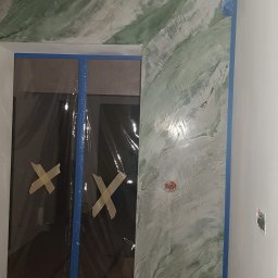 Styloteka - Ściana z dekoracyjnym malowaniem w odcieniach zieleni i szarości, zabezpieczona taśmą malarską, z widocznym fragmentem zakrytego folią ochronną otworu drzwiowego.