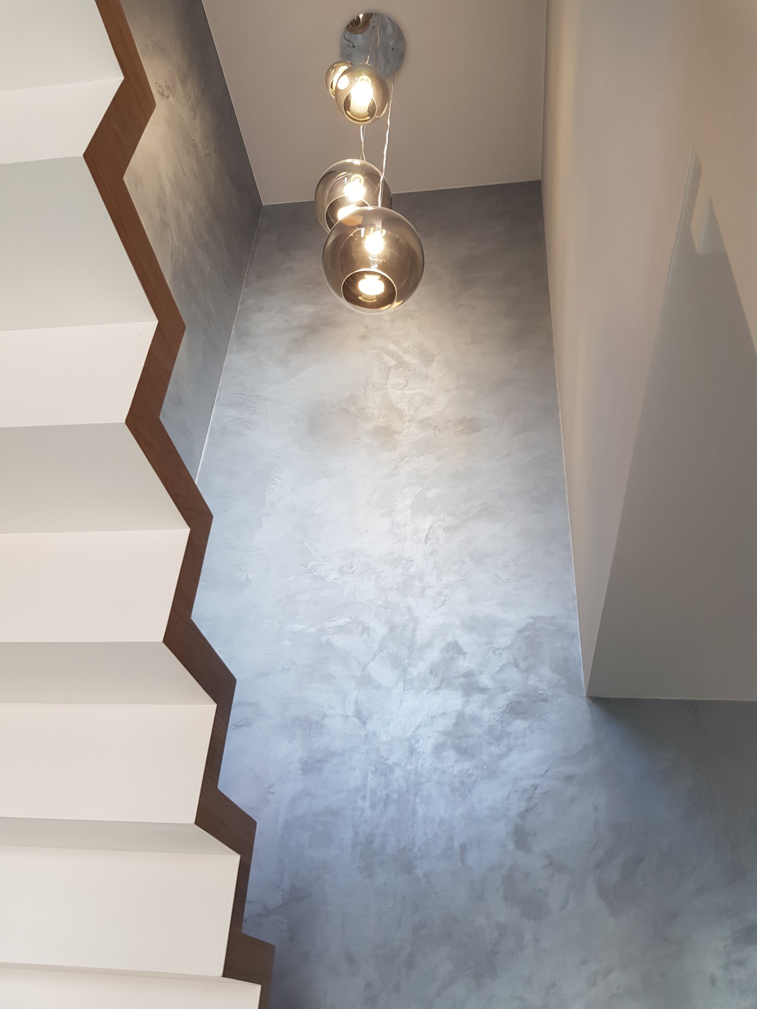 Ściana w odcieniach szarości z efektem dekoracyjnym, oświetlona przez wiszące lampy kuliste, obok białe schody z drewnianymi wstawkami.