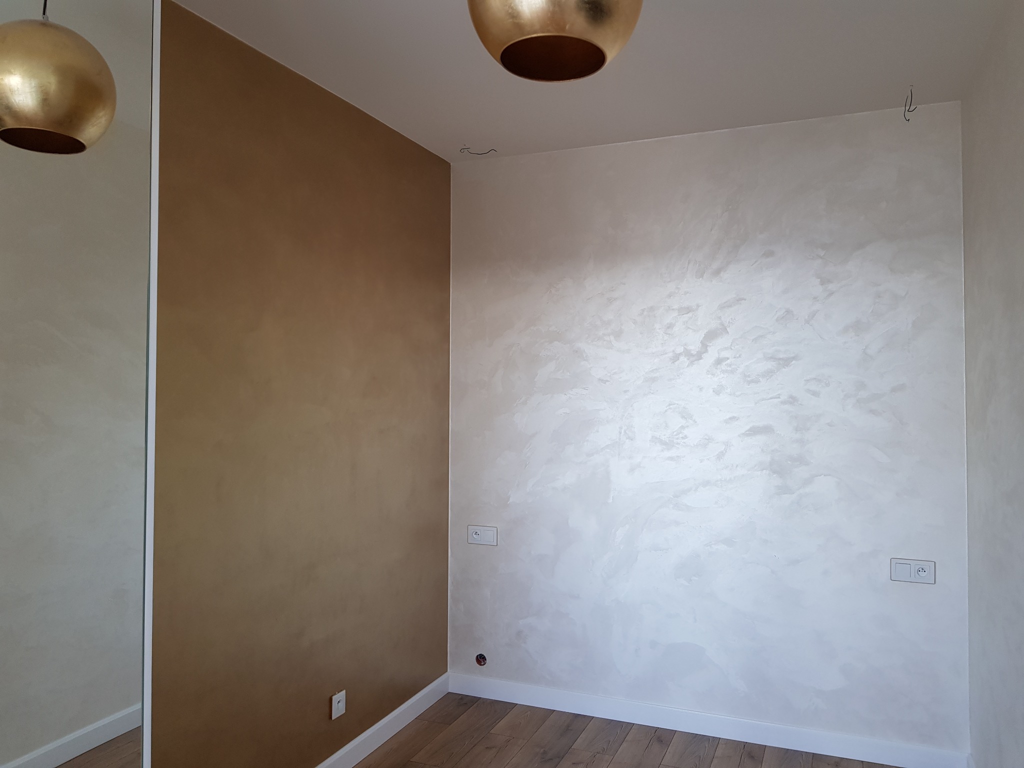 Pokój po malowaniu z efektem dekoracyjnym: jedna ściana w odcieniach brązu, druga w jasnej perle z widoczną fakturą, drewniana podłoga, złota lampa sufitowa.