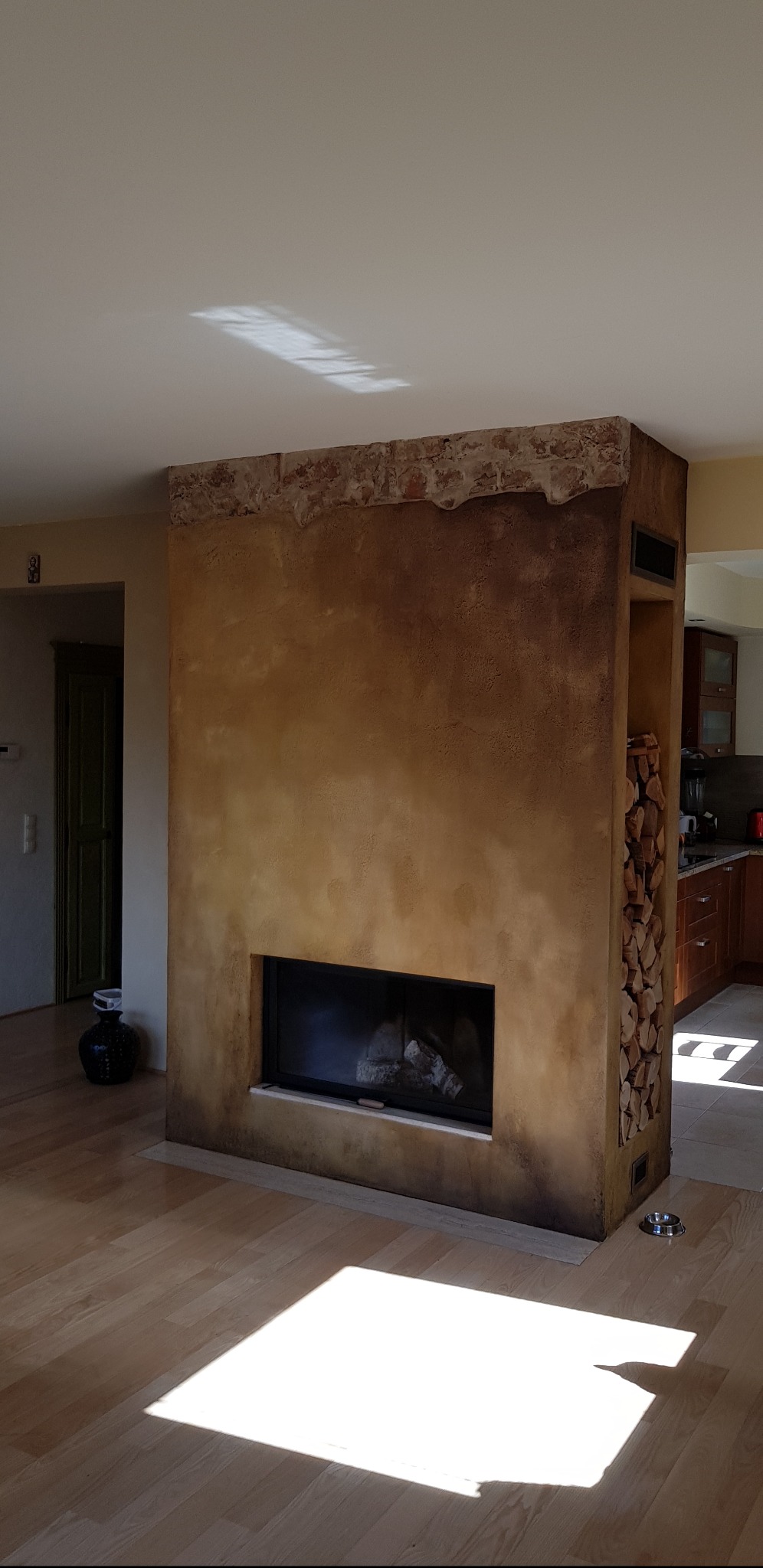 Ściana działowa z kominkiem i wnęką na drewno, wykończona tynkiem dekoracyjnym w odcieniach brązu i beżu, z fragmentem muru z cegły na górze, na podłodze widoczny prostokąt światła słonecznego.