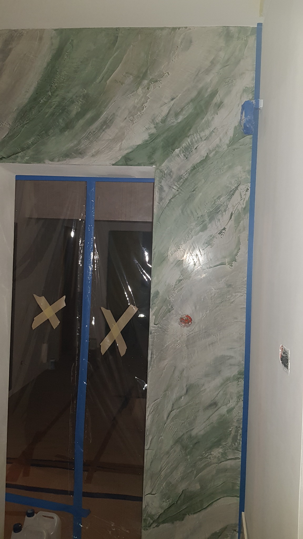 Ściana z dekoracyjnym malowaniem w odcieniach zieleni i szarości, zabezpieczona taśmą malarską, z widocznym fragmentem zakrytego folią ochronną otworu drzwiowego.