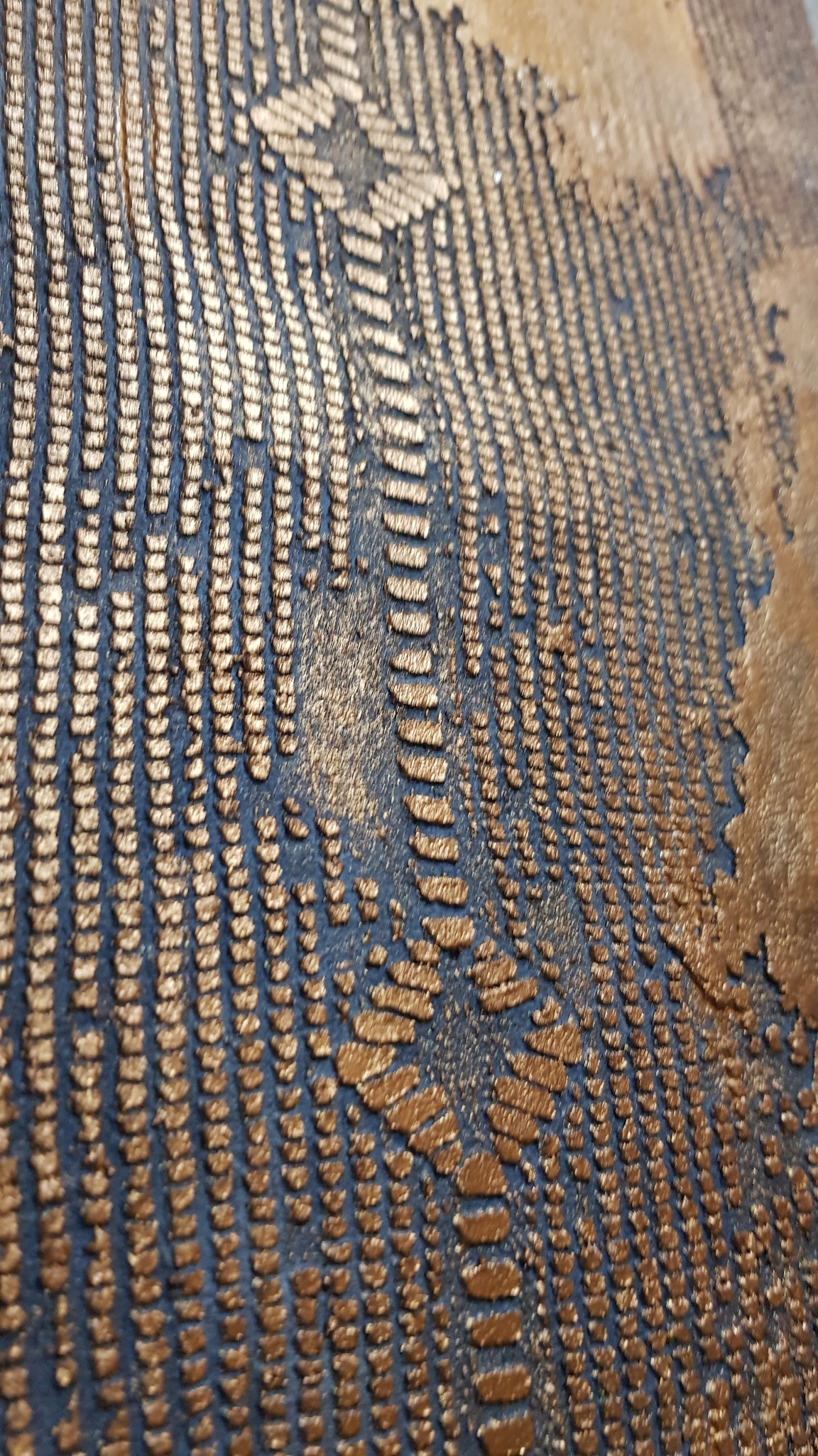 Abstrakcyjna tekstura ściany z metalicznymi, złotymi wypukłościami na ciemnym tle, tworzącymi geometryczny wzór przypominający ślady pędzla.