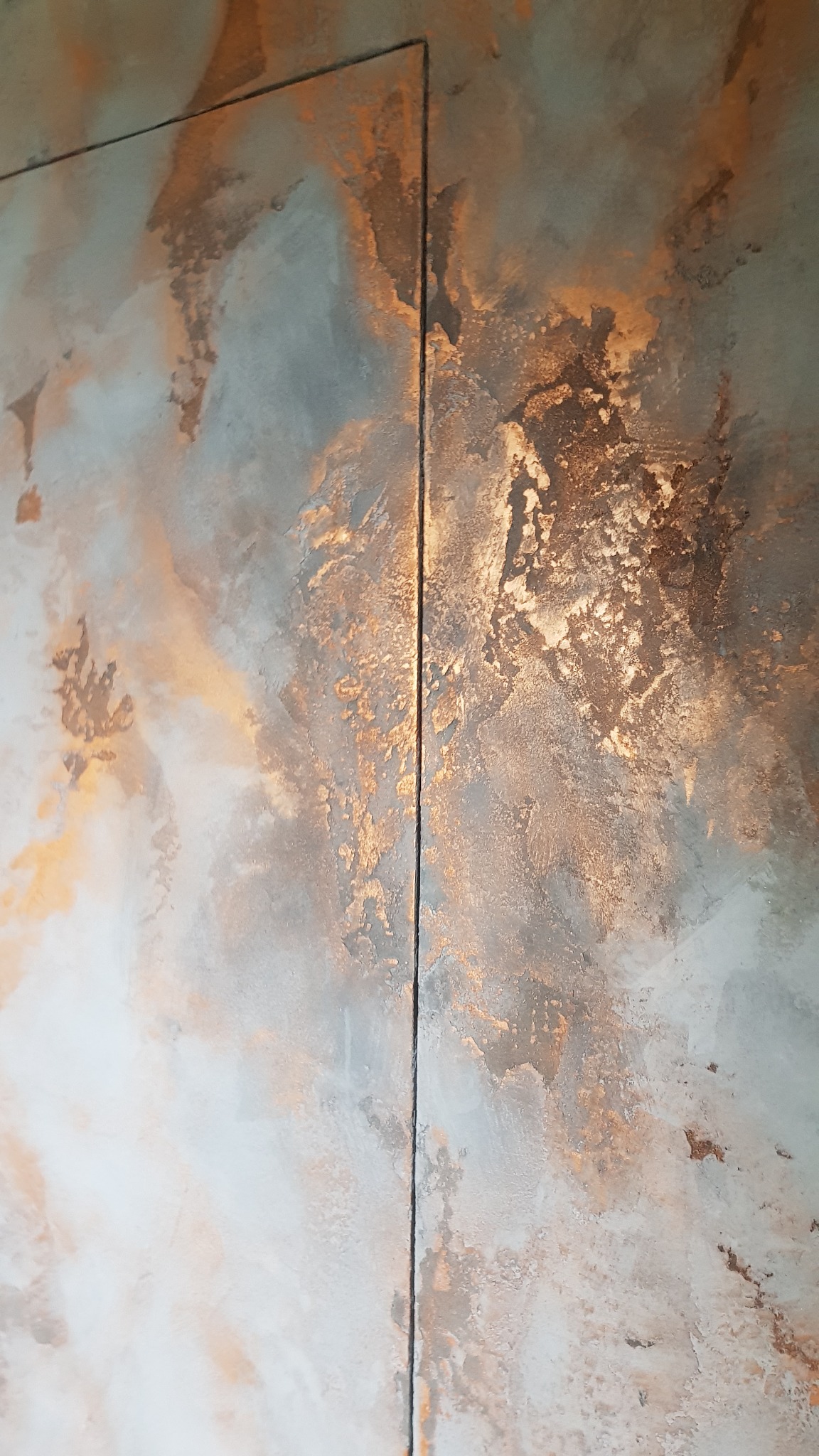 Dekoracyjna ściana z efektem rdzy, łącząca szarości i miedziane akcenty, podzielona pionową linią, ukazująca unikalną teksturę i wzór.