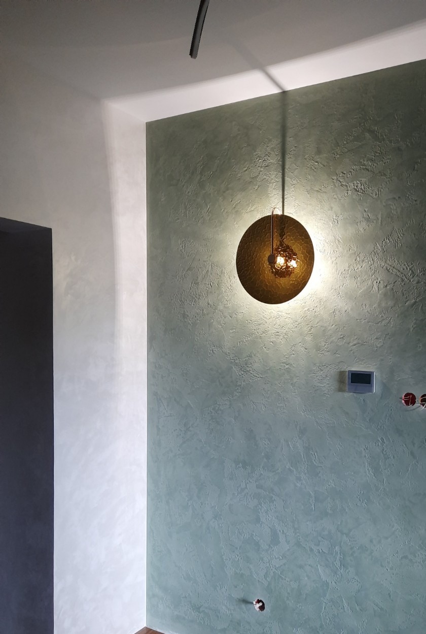 Dekoracyjna ściana w odcieniach zieleni z fakturą w mieszkaniu w Warszawie, oświetlona okrągłą, złotą lampą wiszącą.