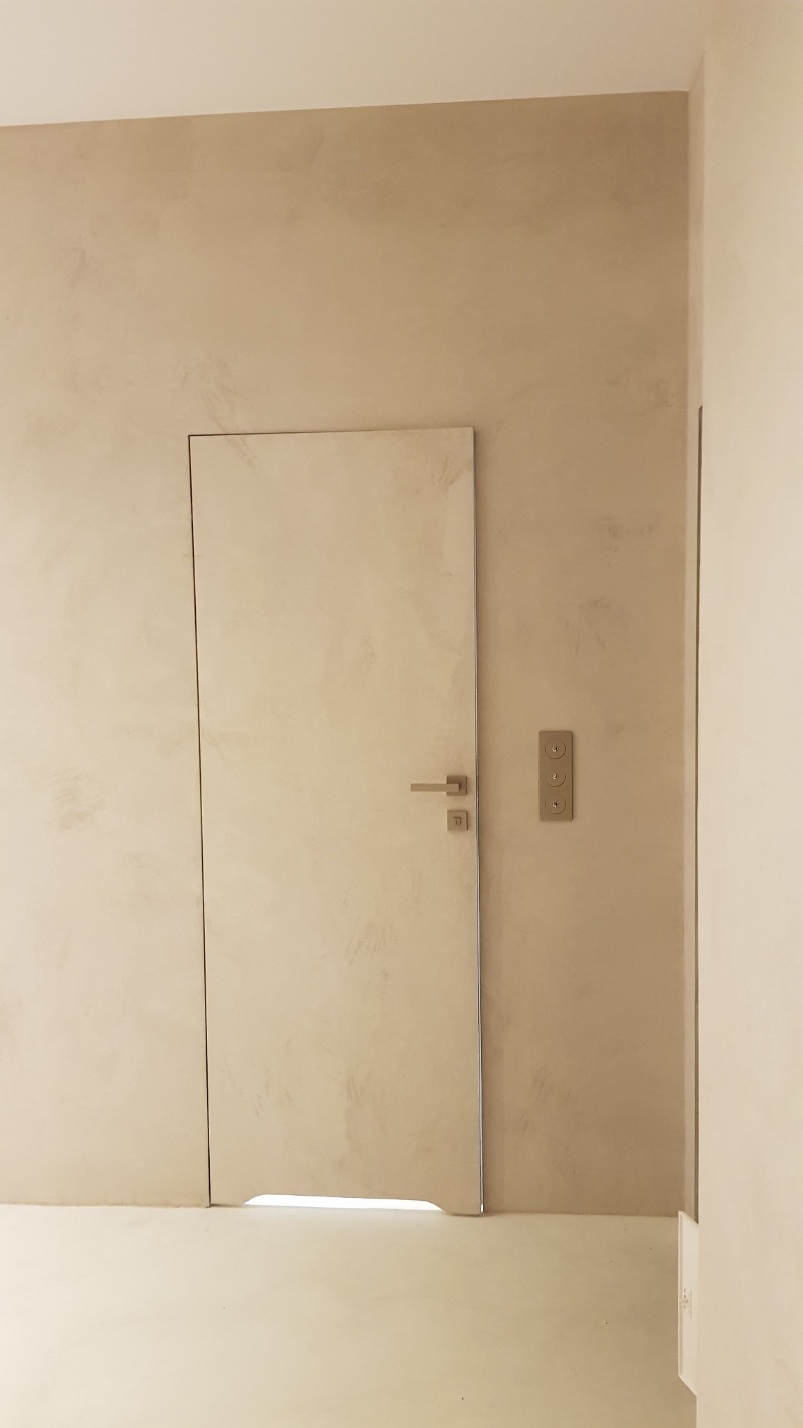 Beżowa ściana z efektem dekoracyjnym, ukryte drzwi z minimalistyczną klamką i panelem sterowania oświetleniem.