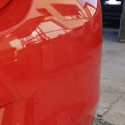 Błyszczący, czerwony błotnik i lampa samochodu po polerowaniu w Grodkowie, z odbiciem budynku w lakierze.