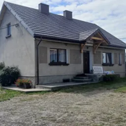 Dom jednorodzinny z nowym dachem z blachodachówki, widoczne kominy, rynny, obróbki blacharskie oraz elementy drewniane więźby dachowej nad wejściem. Na podwórku składowane drewno.