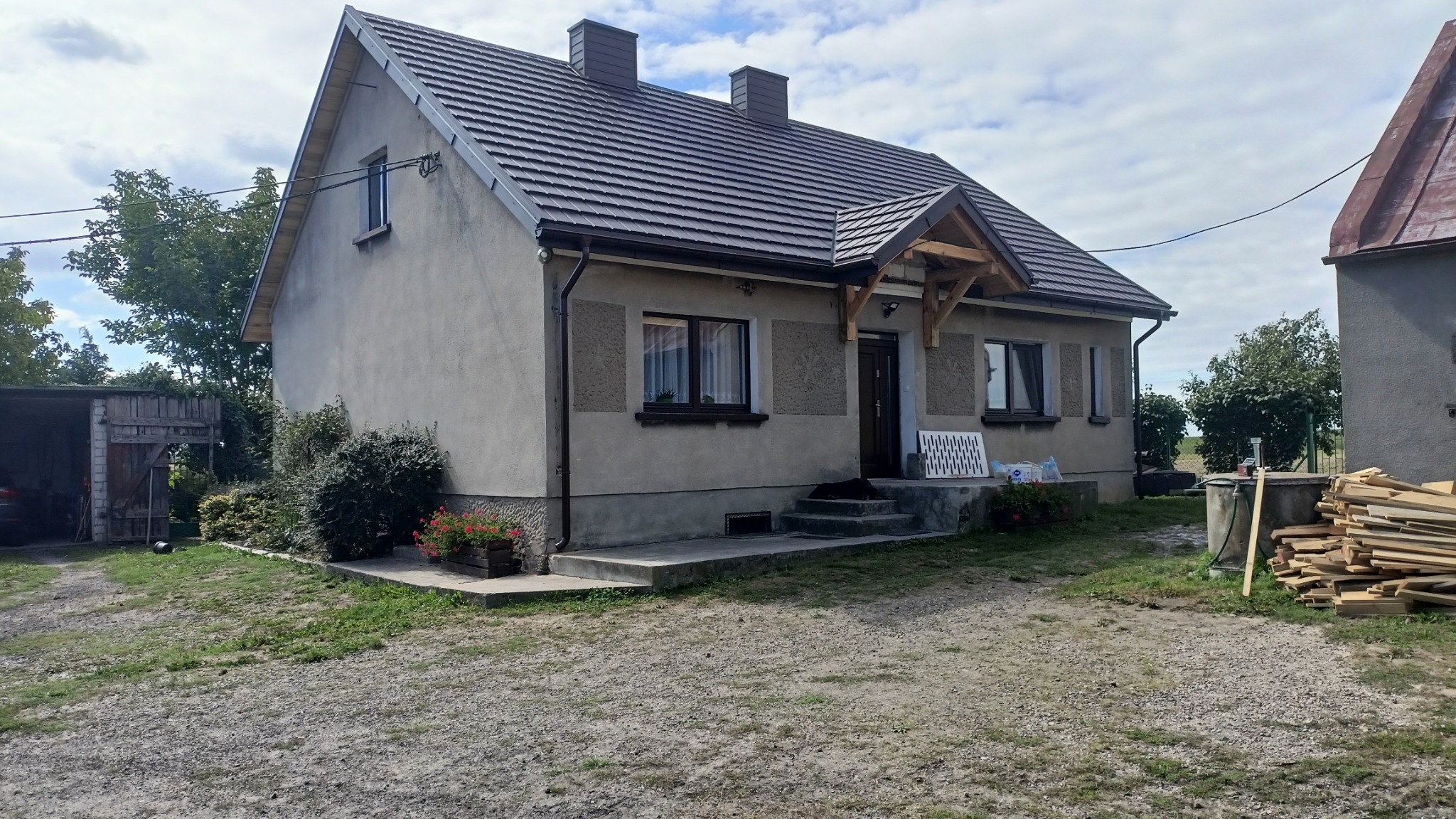 Dom jednorodzinny z nowym dachem z blachodachówki, widoczne kominy, rynny, obróbki blacharskie oraz elementy drewniane więźby dachowej nad wejściem. Na podwórku składowane drewno.