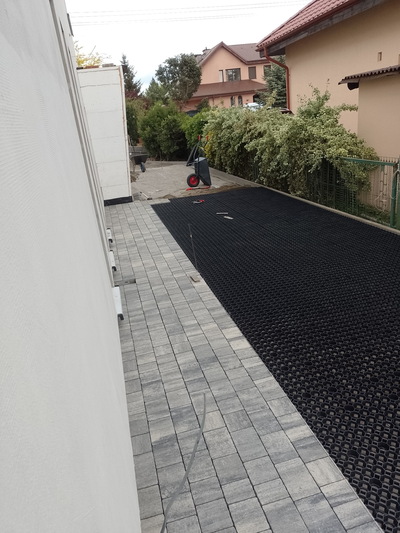 Przygotowanie podłoża pod kostkę: ułożona szara kostka brukowa obok czarnej geokraty, widoczne narzędzia i taczki w tle.