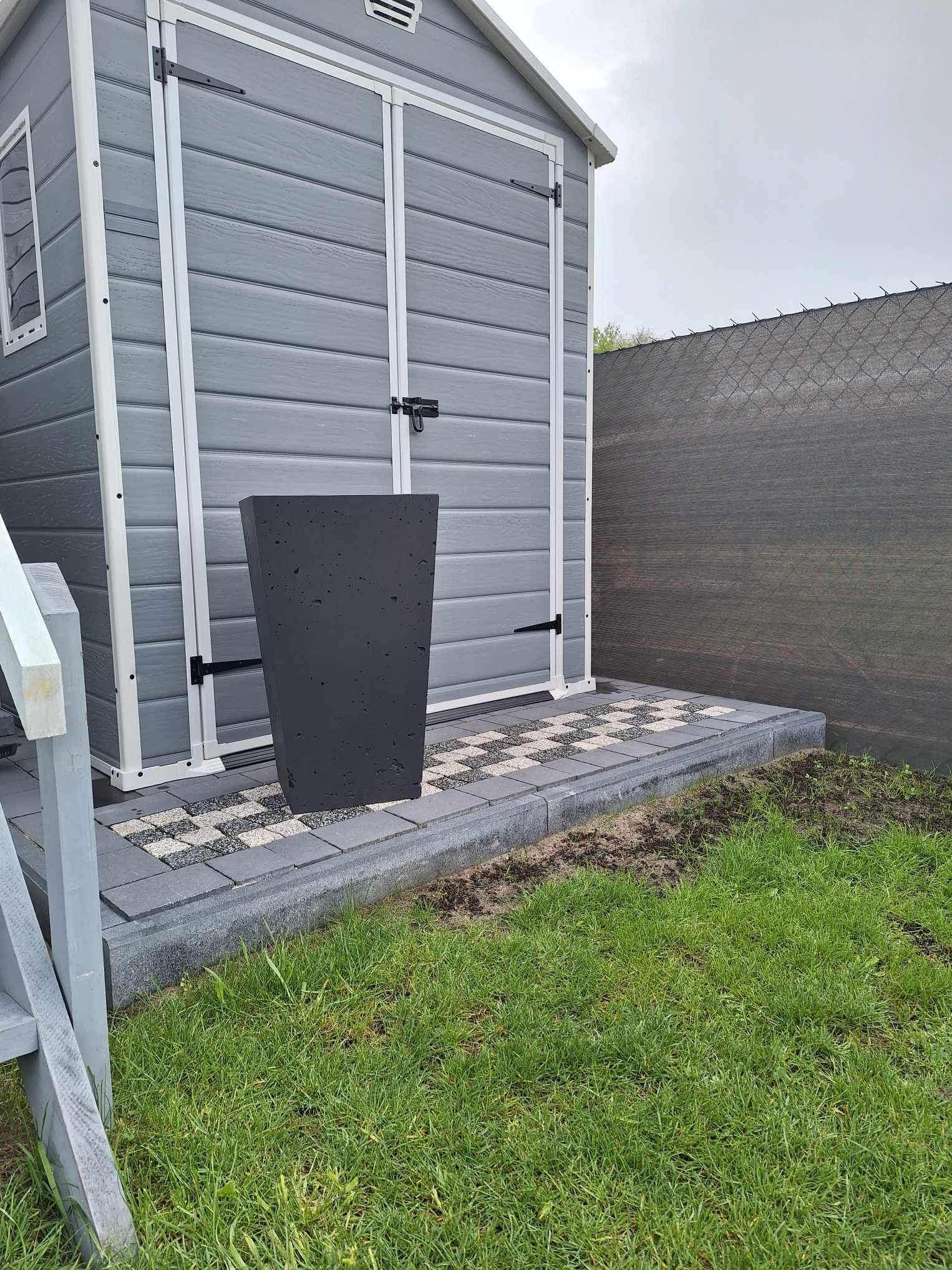 Szara kostka brukowa z czarno-białym wzorem przed szarą szopą ogrodową, z donicą z betonu architektonicznego, świeżo posadzona trawa.