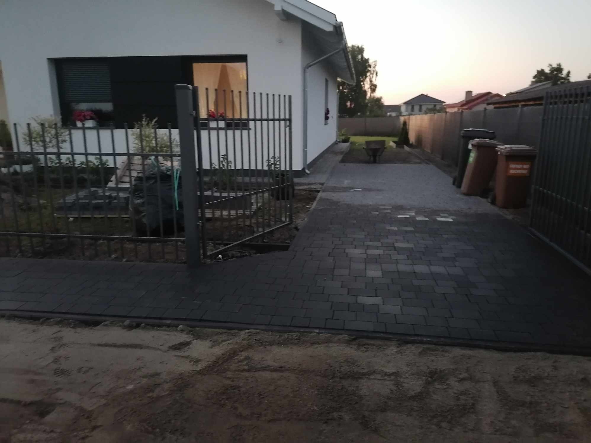 Ułożona, ciemnoszara kostka brukowa prowadząca od furtki do domu jednorodzinnego, widoczne przygotowane podłoże z piasku, w tle kosze na śmieci i sąsiednie zabudowania.