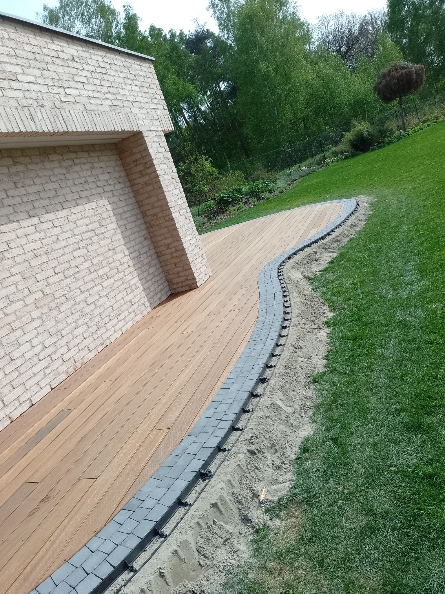 Nowoczesny taras z drewnianą podłogą i obrzeżem z szarej kostki, widoczne czarne elementy mocujące kostkę, jasna ściana budynku z cegły w tle, trawnik i drzewa.