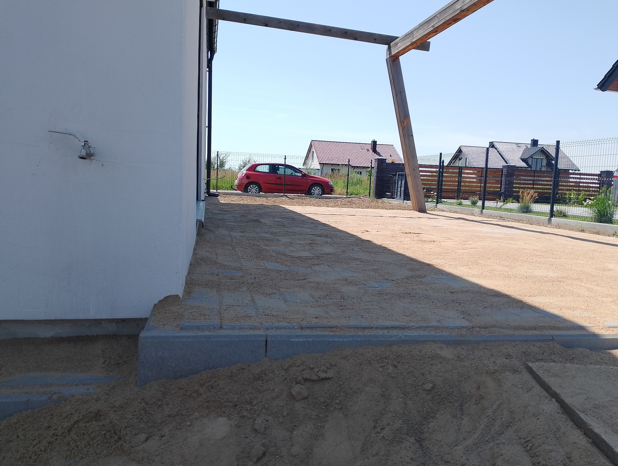 Przygotowany teren pod wiatą, częściowo wyłożony kostką brukową zasypaną piaskiem, widoczny fragment domu z kranem na ścianie i czerwony samochód w tle.