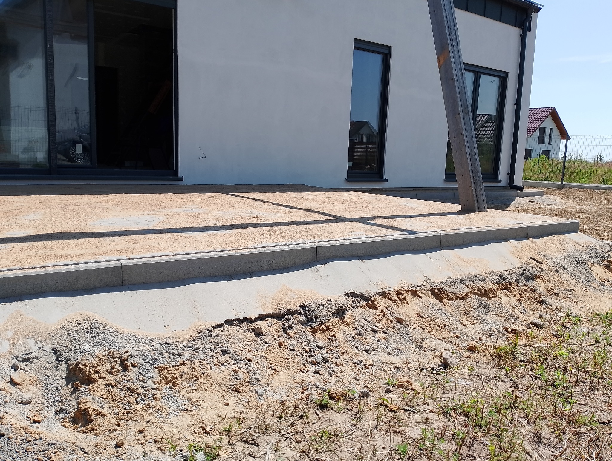 Przygotowanie podłoża pod taras z obrzeżami z kostki betonowej, widoczne piasek, gruz i elementy konstrukcyjne domu w tle.