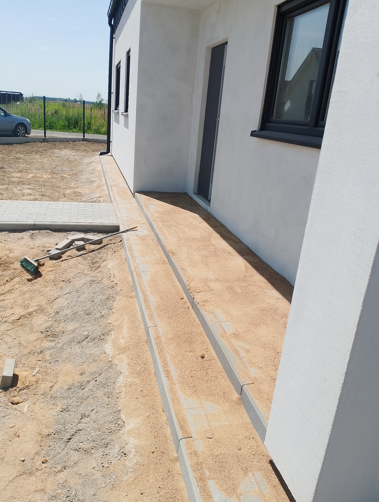 Przygotowanie podłoża z piasku pod ścieżkę z kostki brukowej przy nowym, białym domu z czarnymi oknami; widoczna szczotka brukarska, fragment ułożonej ścieżki i trawnik w tle.