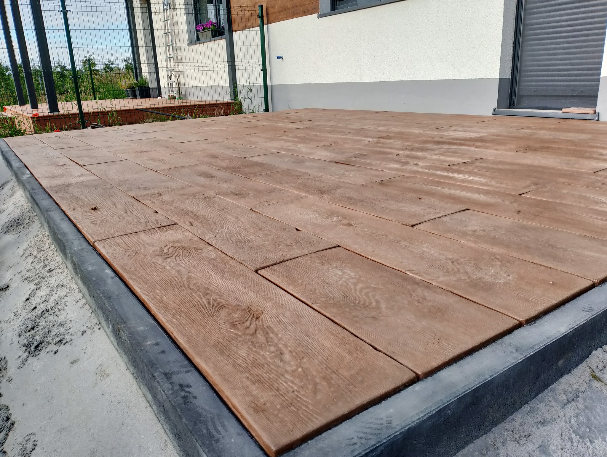 Ułożona kostka brukowa imitująca drewno na tarasie przy domu z widoczną szarą obwódką i fragmentem ogrodu w tle.