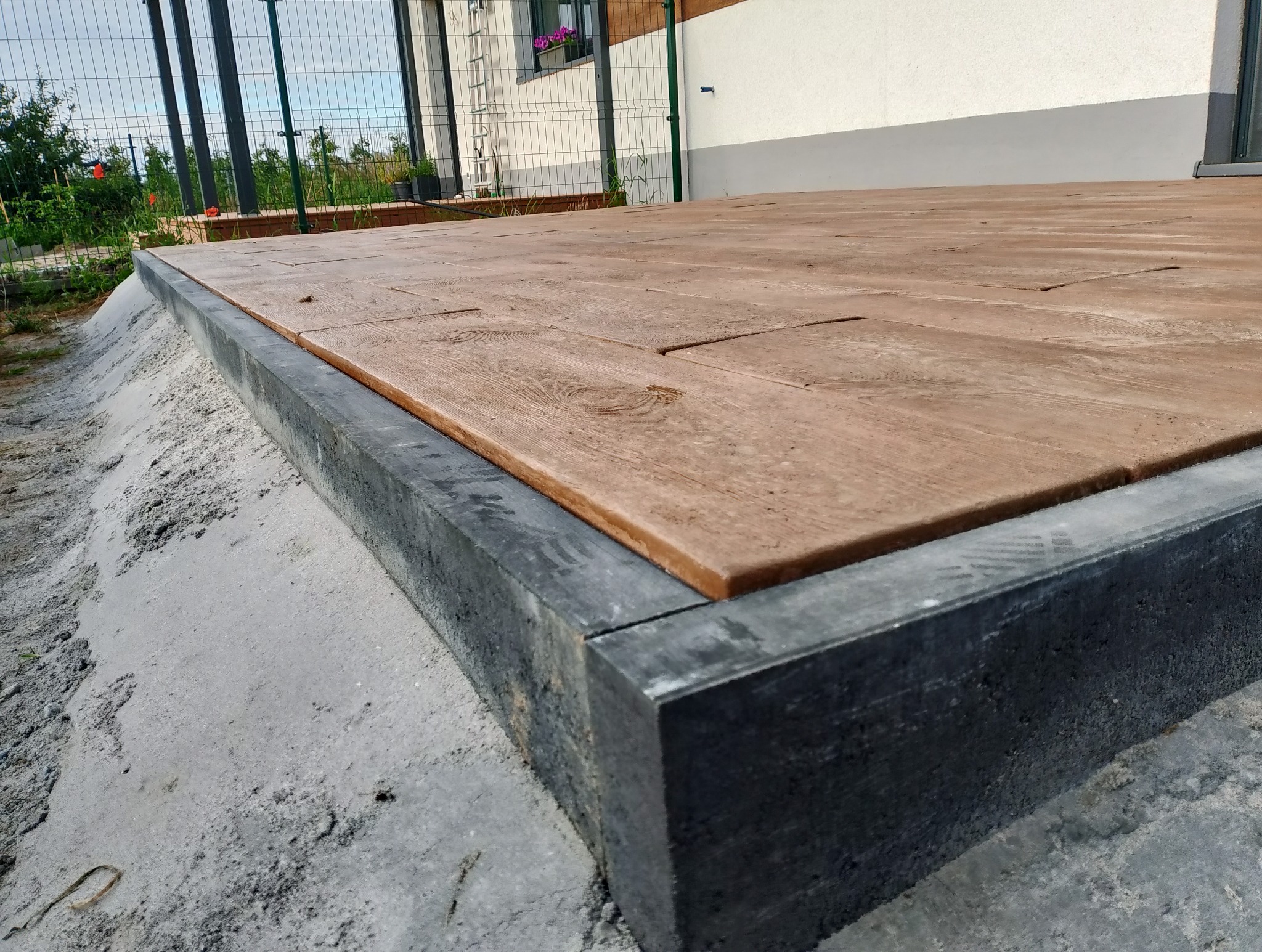 Nowa, brązowa kostka imitująca drewno, obramowana szarą kostką betonową, ułożona na podwórku domu jednorodzinnego, widok z bliska na detale.
