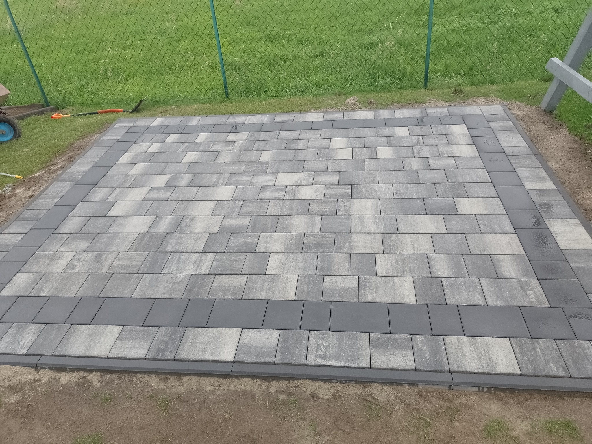 Ułożona prostokątna nawierzchnia z szarej kostki brukowej, obramowana ciemniejszymi, gładkimi elementami, widoczne narzędzia i trawa w tle.