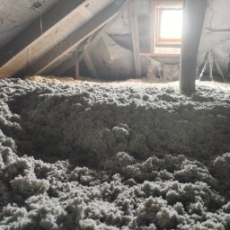 Krukterm Sp. z. o. o - Szara, włóknista izolacja termiczna wypełniająca przestrzeń poddasza z widocznymi elementami drewnianej konstrukcji dachu i małym oknem w tle.