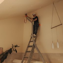 Krukterm Sp. z. o. o - Pracownik na drabinie aplikuje materiał izolacyjny na suficie za pomocą węża, widoczny element oświetlenia i roślina w tle.